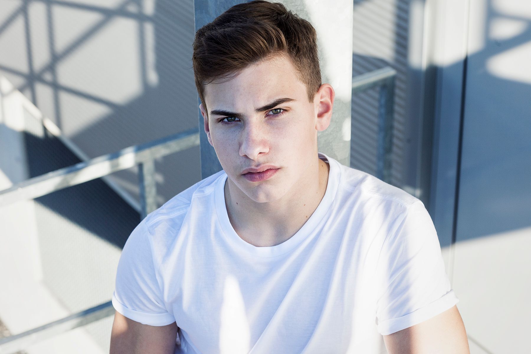portrait; young guy in white shirt sitting on a stairway; light blue background