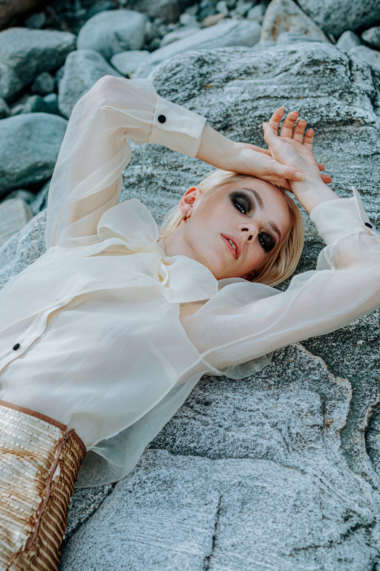 blonde girl with dark eye makeup; golden skirt, laying on stone outside